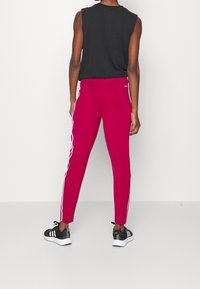 Figurbetonte rosa Sporthose mit weißen Seitestreifen, kombiniert mit einem schwarzen ärmellosen Oberteil und schwarzen Sportschuhen. Glatte Textur.