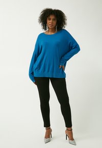 Blauer übergroßer Pullover mit langen Ärmeln, strukturiertem Stoff und lockerer Passform, kombiniert mit schwarzen eng anliegenden Hosen und glitzernden High Heels.