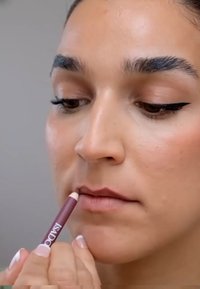 Läppens penna i en mauve nyans hålls nära läpparna. Huden har en jämn textur med en naturlig ton. Fin eyeliner framhäver ögonen. Neutral bakgrund.