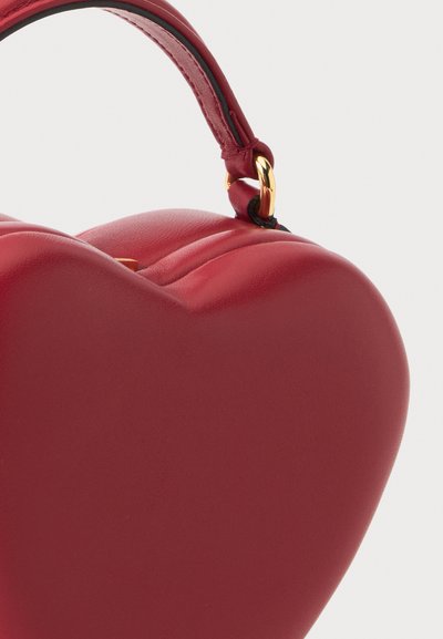 Sac à main rouge en forme de cœur avec une courte bandoulière attachée par un anneau métallique doré sur un fond clair.