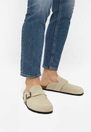 Sabots en cuir suédé beige avec un bout rond, dotés d'une sangle unique et d'un détail de boucle. Portés avec un jean en jean bleu. Semelle plate noire.