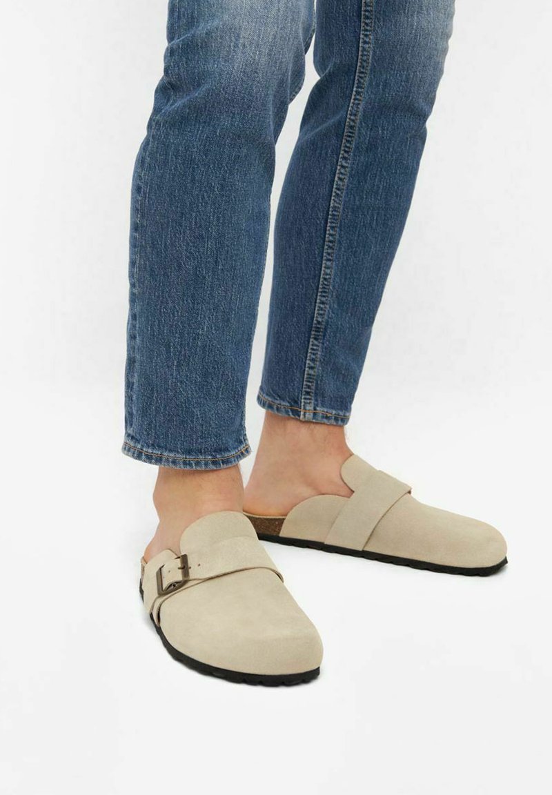 Zoccoli in suede beige con punta rotonda, dotati di una singola cinghia e dettaglio della fibbia. Indossati con jeans di denim blu. Suola piatta nera.
