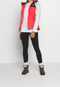 Czerwona i szara wodoodporna kurtka, biały t-shirt, czarne legginsy oraz ciemnozielone buty trekkingowe. Teksturowane materiały z widocznymi detalami metalowymi.