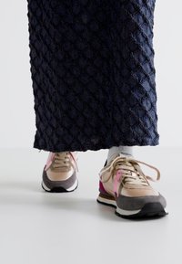 Baskets avec une tige beige et grise, des accents roses, un design texturé et une semelle blanche. Portées avec une jupe noire à motifs.