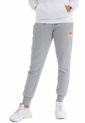 Person iført grå ellesse joggingbukser, hvid sweatshirt og hvide sneakers, stående med hænderne i sweatshirtlommen.