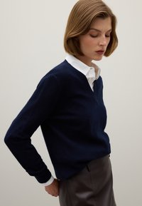 Marineblauwe wollen trui met een V-hals ontwerp, gecombineerd met een wit overhemd met knopen en kraag, tegen een neutrale achtergrond.