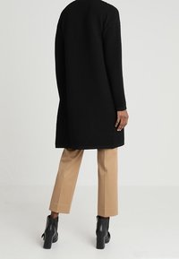 Svart ribbad lång kappa med en avslappnad passform, i kombination med beige tapered byxor och svarta ankelstövlar, som visar upp ett texturerat tyg.