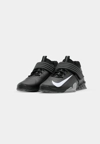 Chaussures de sport noires avec des surfaces texturées, un tissu en maille et des fermetures à double sangle. Présente un grand logo Nike en blanc sur le côté.