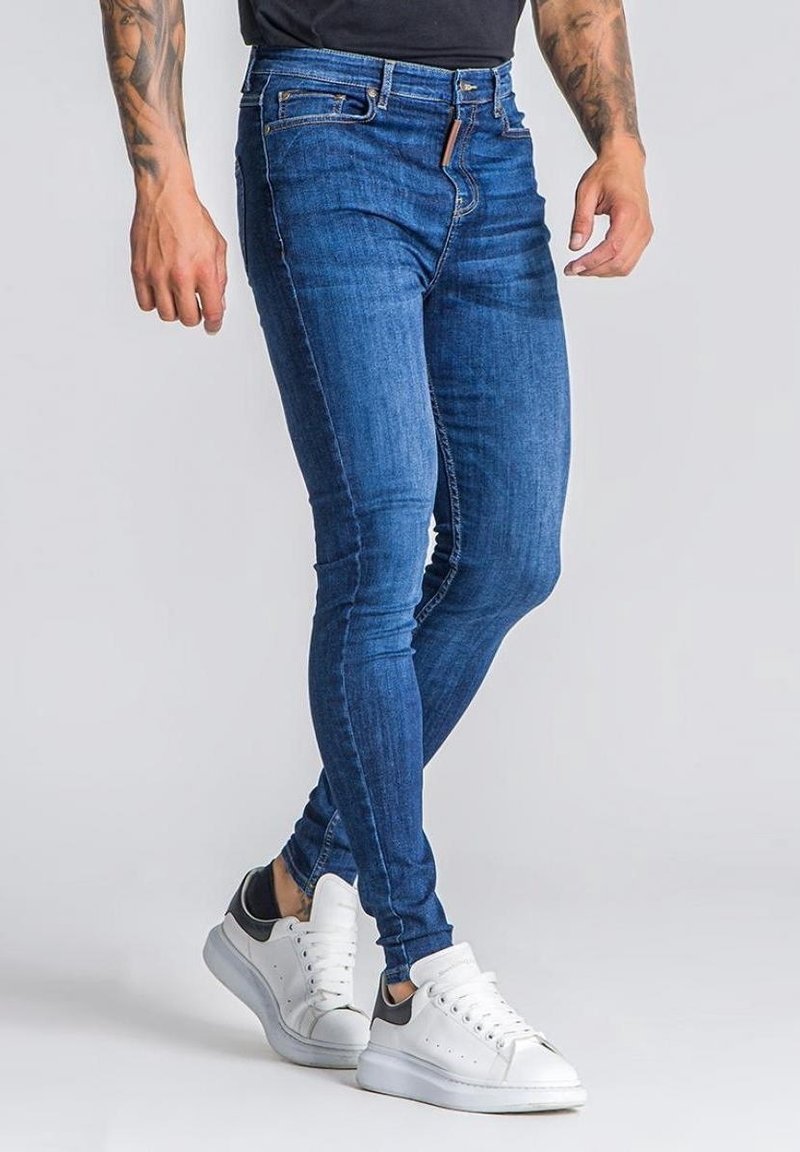 Vaqueros ajustados en denim azul oscuro, con un diseño clásico de cinco bolsillos y un ligero desvanecimiento, combinados con zapatillas blancas con detalles en gris.