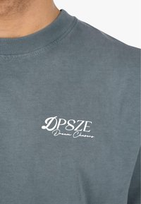 Grå bomull t-skjorte med rund hals, med en hvit logotype som leser "DPSZE Dream Chasers" på venstre brystområde.