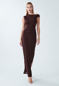 Bruine, nauwsluitende avondjurk met geplooide lijfje en off-shoulder ontwerp. Soepele textuur, tot op de grond, met een zijslit en minimale accenten.