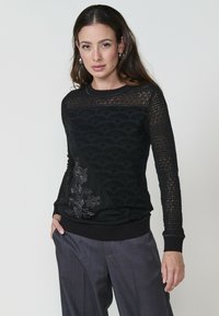 Donna con lunghi capelli scuri, che indossa un maglione nero con un motivo in pizzo e decorazioni floreali, abbinato a pantaloni grigi, in posa con sicurezza.