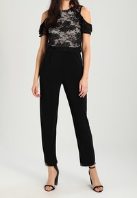Czarny kombinezon z koronkowym gorsetem i odkrytymi ramionami. Spodnie są dopasowane o prostym kroju. Uzupełniony o wysokie obcasy.