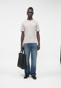 Tricou gri deschis, blugi din denim albastru, pantofi din piele negri și o geantă tote neagră. Modelul stă în fața unui fundal alb.