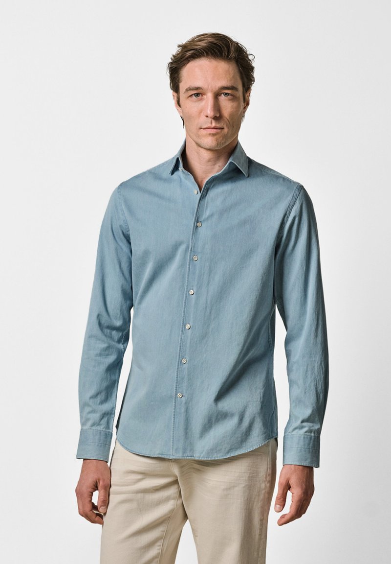 Homme aux cheveux bruns courts portant une chemise bleu clair à manches longues boutonnée et un pantalon beige, debout devant un fond blanc uni.