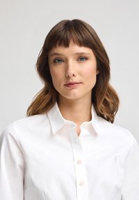 Camisa blanca de botones con cuello clásico. Presenta un sutil logotipo en el pecho y una textura suave y pulida.