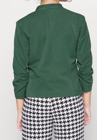 Zelený přizpůsobený sako s nařasenými rukávy, hladkou texturou a jednoduchým límcem, v kombinaci s kalhotami s černo-bílým vzorem kožešiny.