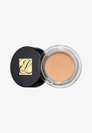 Fond de teint crémeux dans un pot en verre transparent ; le couvercle noir est orné d'un logo doré. Couleur beige clair avec une texture lisse.