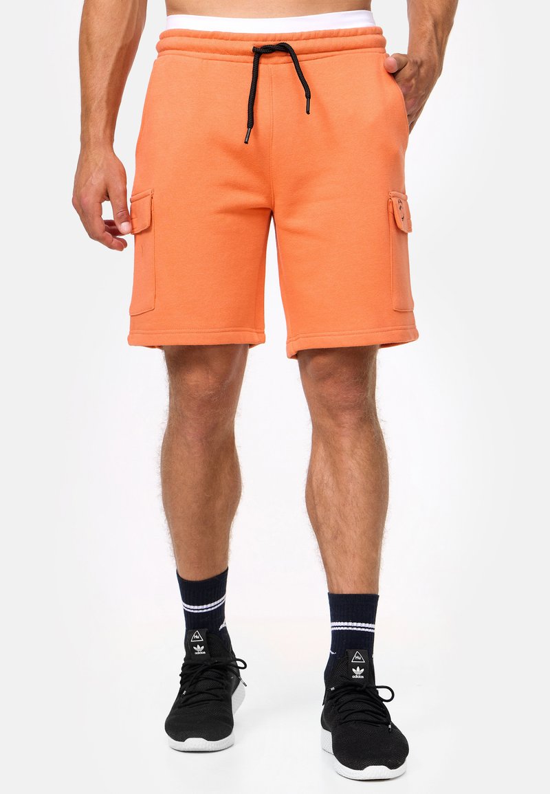 Shorts en coton orange avec poches latérales, taille élastique et cordon noir. Associés avec des chaussures noires et des chaussettes rayées bleu marine.
