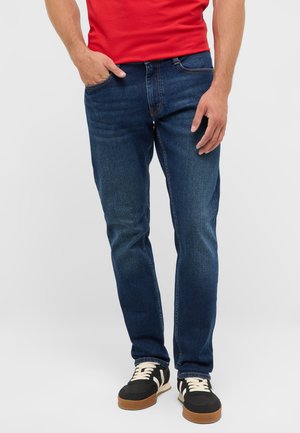 Homme portant un jean bleu foncé, des baskets noires avec des lacets et semelles blanches, et une chemise rouge, debout avec une main dans la poche.