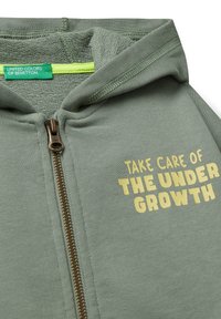 Groene rits hoodie met "Zorg voor de ondergroei" in geel gedrukt op de borst en een label van "United Colors of Benetton" binnenin.