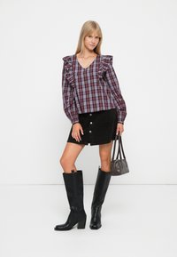 Geruite blouse met gerimpelde schouders in bordeaux en blauw, gecombineerd met een zwarte corduroy rok met knopen en kniehoge zwarte laarzen.