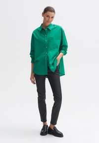 Groene langemouwige overhemden met knopen en een borstzak, gedragen over zwarte slim-fit broeken, gecombineerd met zwarte loafers. Soepele katoenen textuur.