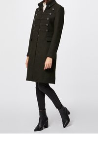 Donkergroen wollen jas met een hoge kraag, voorzien van opvallende knoopdetails en een rechte snit. Gecombineerd met zwarte jeans en enkellaarzen.