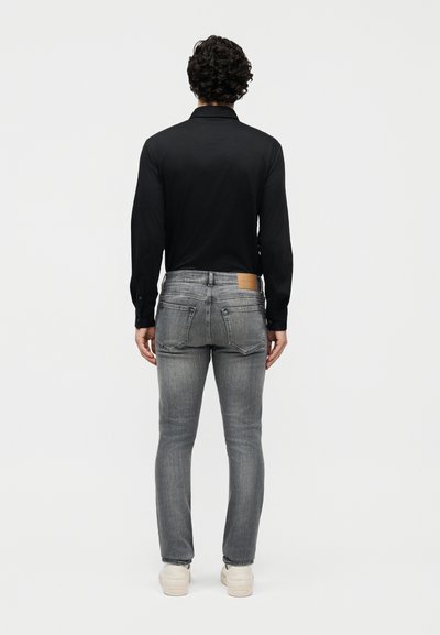 Άνδρας με κατσαρά μαλλιά που φοράει μαύρο πουκάμισο με μακριά μανίκια, γκρι στενά τζιν και λευκά αθλητικά παπούτσια, στέκεται με την πλάτη προς το μέρος μας σε απλό φόντο.