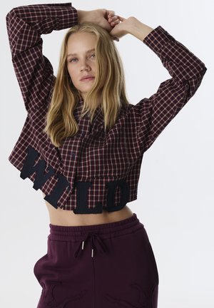 Camicia a quadretti tagliata in borgogna e blu navy, con bottoni blu e scritta 'WILD' sull'orlo, abbinata a pantaloni sportivi viola.