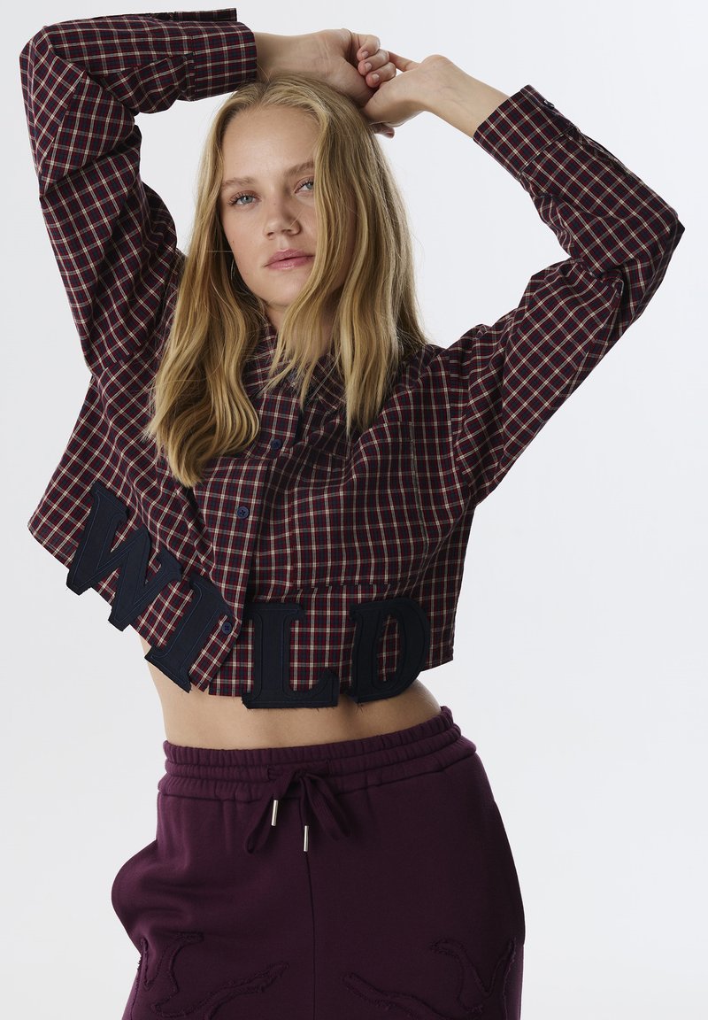 Geknipte geruite shirt in bordeaux en marineblauw, met blauwe knopen en 'WILD' opschrift op de zoom, gecombineerd met paarse joggingbroek.