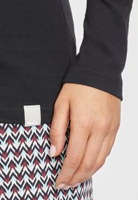 Schwarzes Langarmshirt mit gerippter Textur, das an dem Saum mit einem Bench-Logo versehen ist. Gezeigt mit gemustertem, mehrfarbigem Stoff darunter.