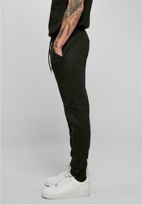 Pantalon de jogging vert foncé avec une coupe slim, doté de poches latérales et d'une taille élastique avec cordon de serrage, associé à des baskets blanches.