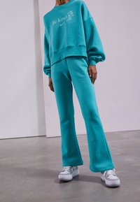 Teal cropped sweatshirt with "Be kind" text, paired with matching flared sweatpants; both made of soft fabric. White shoes complete the outfit.