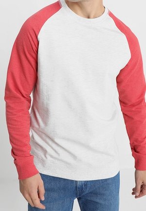 Hombre con camisa blanca de manga larga con mangas raglán rojas y vaqueros azules, de pie contra un fondo blanco liso.