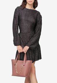 Rosa læder tote taske med kædeaccent, rektangulær form og dobbelte håndtag, holdt foran en sort polka dot kjole.