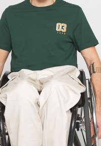 Zelené bavlněné tričko s krátkým rukávem, na levé straně hrudi s bílou a oranžovou grafikou "03 TEAM". Viditelné béžové cargo kalhoty.