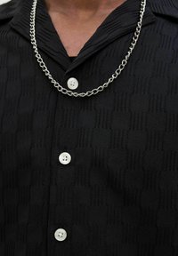 Chemise noire texturée à boutons avec des motifs horizontaux et verticaux, boutons blancs et collier en chaîne argentée.