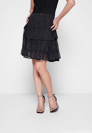 Femme portant une jupe noire à pois en couches et des sandales noires à talons hauts avec des brides en strass, posant les mains sur les hanches.