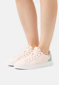 Persona che indossa sneakers rosa chiaro allacciate con suole bianche e dettagli verdi sul tallone, in piedi su una superficie bianca.