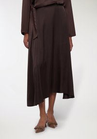 Gonna di satin marrone con orlo asimmetrico, legata in vita con una cintura coordinata. Abbinata a scarpe slingback marroni con dettagli a fiocco.