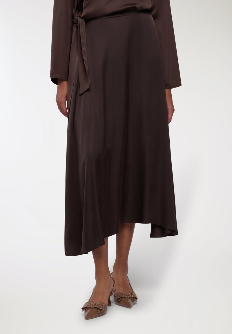 Gonna di satin marrone con orlo asimmetrico, legata in vita con una cintura coordinata. Abbinata a scarpe slingback marroni con dettagli a fiocco.
