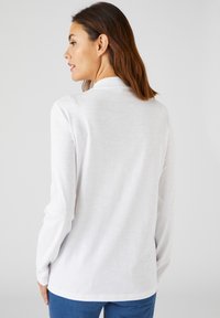 Witte langmouwen shirt met een gladde textuur en een hoge kraag, gecombineerd met blauwe jeans. De achterkant laat een ontspannen pasvorm en strakke lijnen zien.