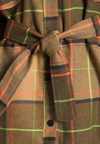 Brązowa tkanina w kratę z pomarańczowymi, zielonymi i niebieskimi liniami. Zawiera związany pasek w talii i detal z guzikami. Miękka struktura o ciepłym wyglądzie.