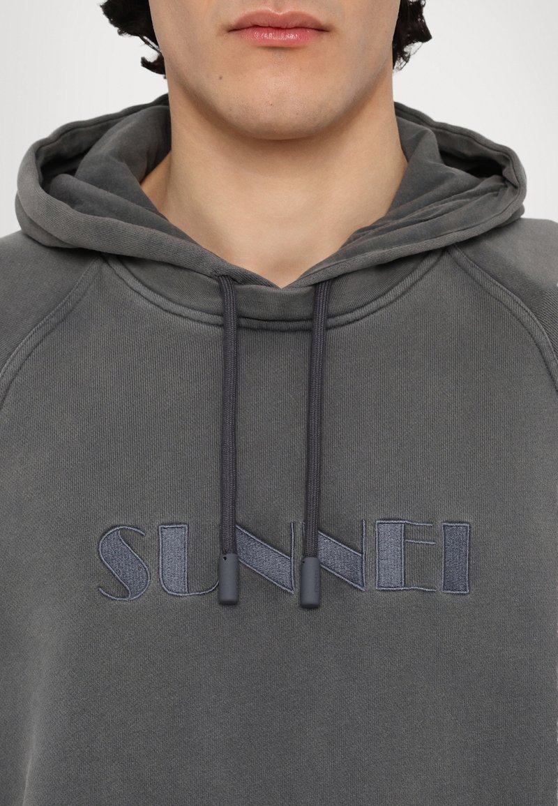 Haut à capuche gris avec poche kangourou, cordons de serrage avec embouts en caoutchouc, et logo "SUNNEI" brodé en gris foncé. Tissu doux et texturé.
