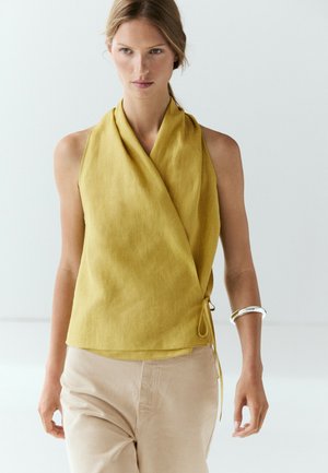 Femme portant un haut cache-cœur sans manches moutarde et un pantalon beige avec un bracelet argenté au poignet gauche sur fond blanc.