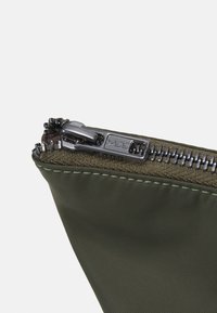 Gros plan sur une pochette en tissu vert foncé partiellement ouverte avec une fermeture éclair en métal noir et des coutures visibles.