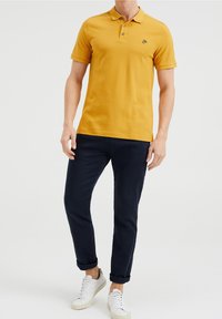 Kortärmad gul polo med tre knappar, i kombination med marinblå byxor och vita sneakers. Har en liten broderad logotyp på bröstet.