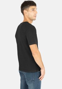 Schwarzes T-Shirt mit kurzen Ärmeln und Rundhalsausschnitt, aus weicher Baumwolle, seitlich präsentiert, kombiniert mit blauen strukturierten Jeans.