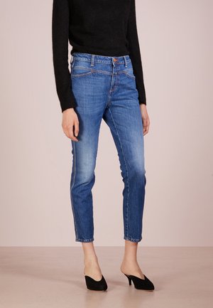 Jean en denim bleu clair avec une coupe fuselée, taille mi-haute et un léger délavage. Comprend cinq poches et des accents de couture. Associé à des talons noirs.